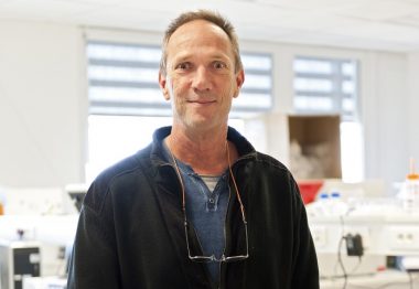 Le professeur debout devant uine paillasse de laboratoire.