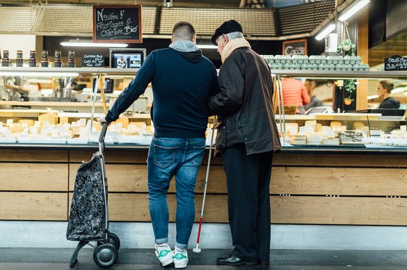 Un auxiliaire de vie accompagne un vieux monsieur aveugle devant un étal de fromages pour faire des courses