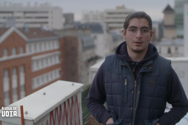 Idriss, avec luettes et cheveux attachés sur un balcon de face